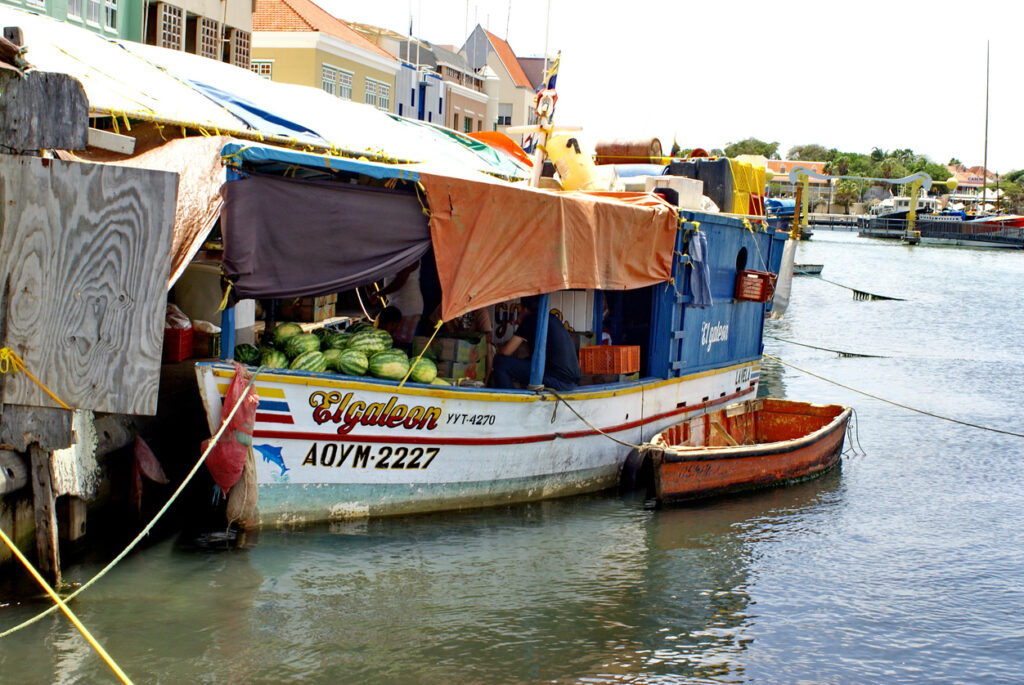 Colorful produce boat at the Floating Market in Willemstad, Curaçao, with watermelons stacked at the bow and vendors working under shaded tarps along the waterfront