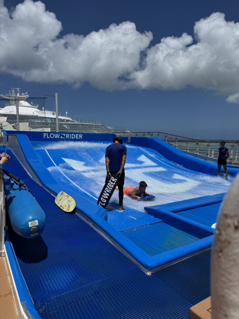 Instructor assisting a guest on the FlowRider surf simulator aboard a cruise ship, with water rushing across the wave surface under a bright sky.