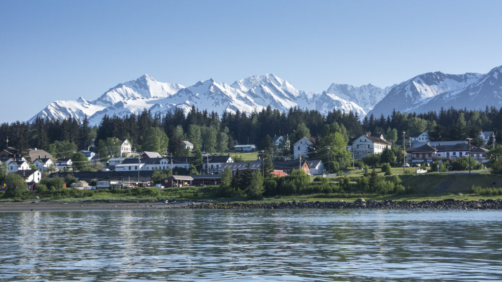 A scenic view of a small Alaskan town along the waterfront, surrounded by dense evergreens and backed by towering snow‑covered mountains under a clear blue sky.
