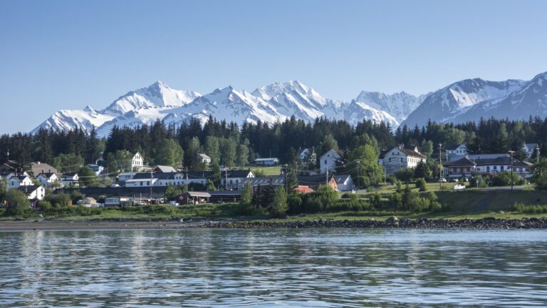A scenic view of a small Alaskan town along the waterfront, surrounded by dense evergreens and backed by towering snow‑covered mountains under a clear blue sky.