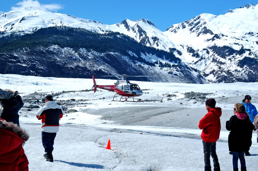 Red helicopter landed on a snowy glacier in Juneau, Alaska, with visitors standing nearby against rugged mountain scenery.