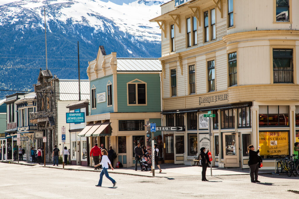 Broadway Street in downtown Skagway, Alaska, with shoppers walking past restored Gold Rush–era false‑front buildings, wooden boardwalks, and local shops on a spring day.