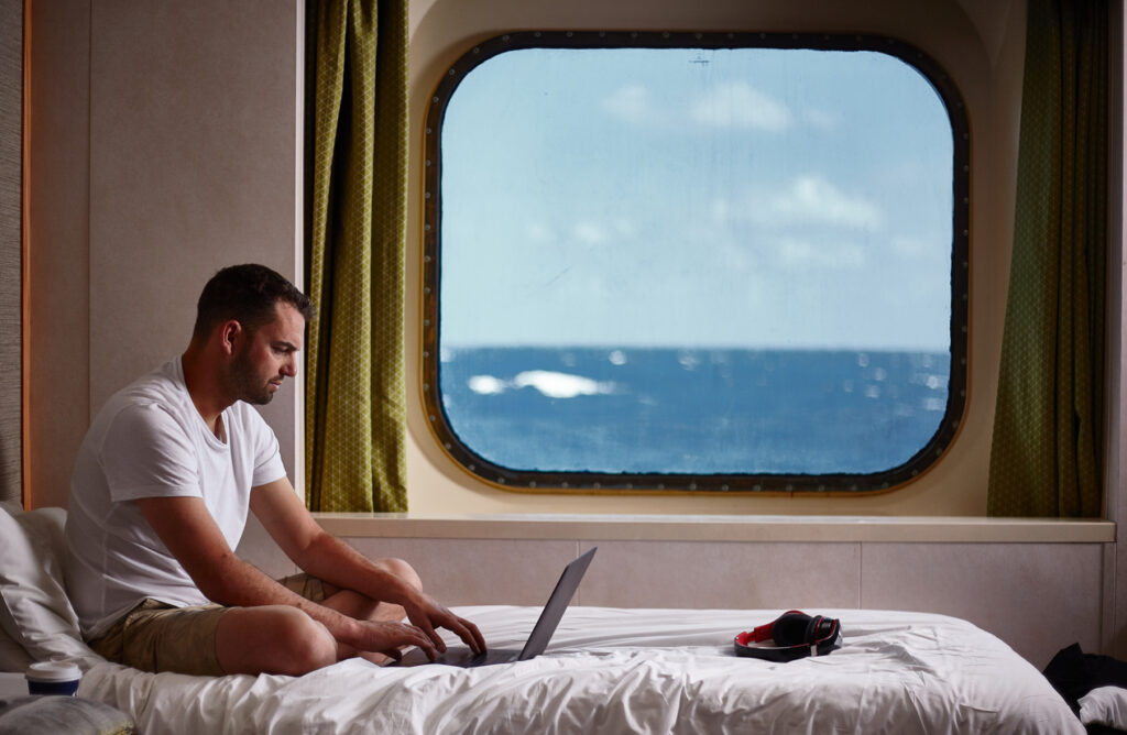 Person working on a laptop in a cruise cabin overlooking the ocean, illustrating how cruise wifi works for staying connected at sea
