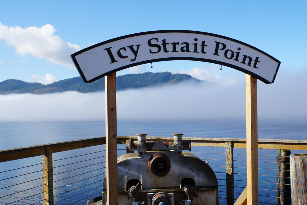 A waterfront view of Icy Strait Point with forested hills, calm water, and distant mountains under a clear sky.