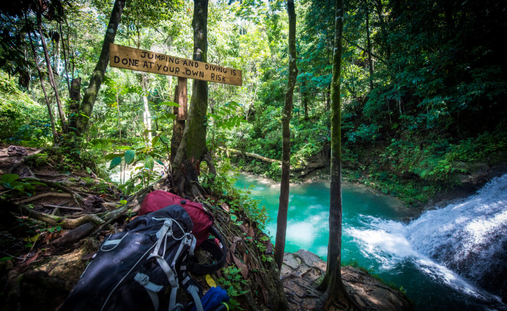 Backpacks resting on the ground beside a “Jumping and diving is done at your own risk” sign at Irie Blue Hole in Ocho Rios, with a waterfall, turquoise water, and tropical forest in the background.