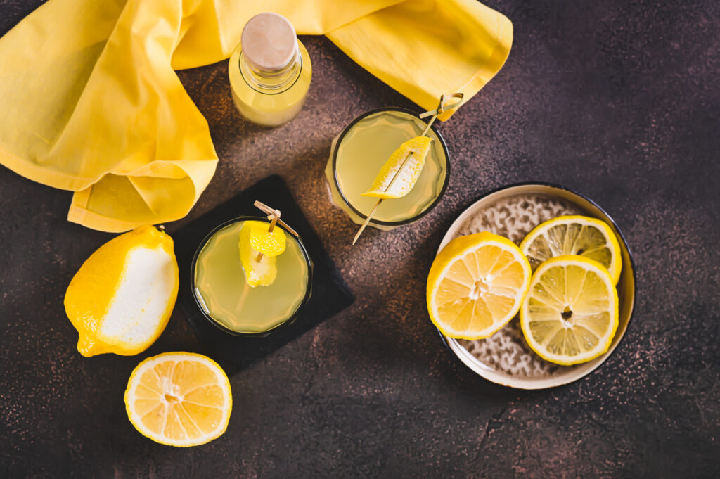 Glasses and a small bottle of bright yellow limoncello surrounded by fresh lemons and lemon peels on a dark surface