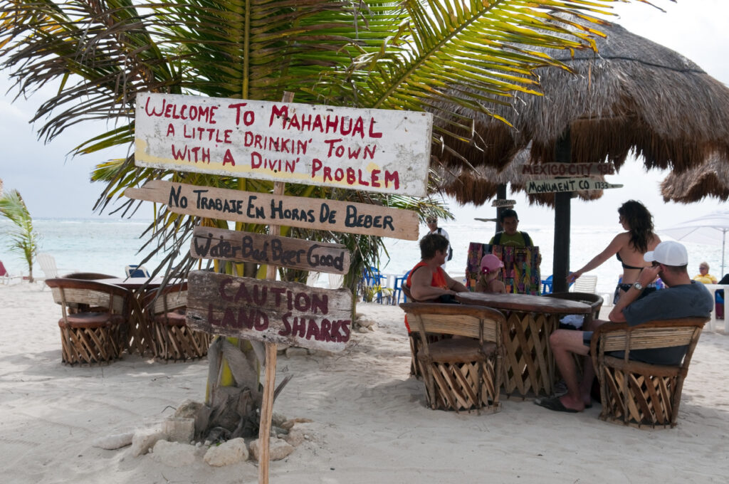 Beachside café scene in Mahahual, Costa Maya, with tourists seated at a table while a local vendor displays handmade bracelets, and humorous wooden signs adding to the relaxed, tropical atmosphere.