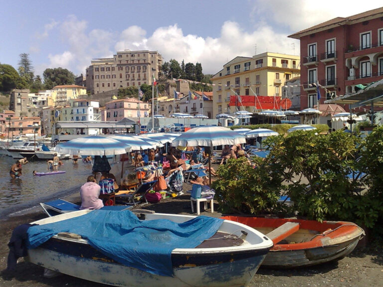 Busy beach at Marina Piccola in Sorrento with blue‑and‑white umbrellas, lounge chairs, small boats on the shore, and colorful buildings rising along the cliffs