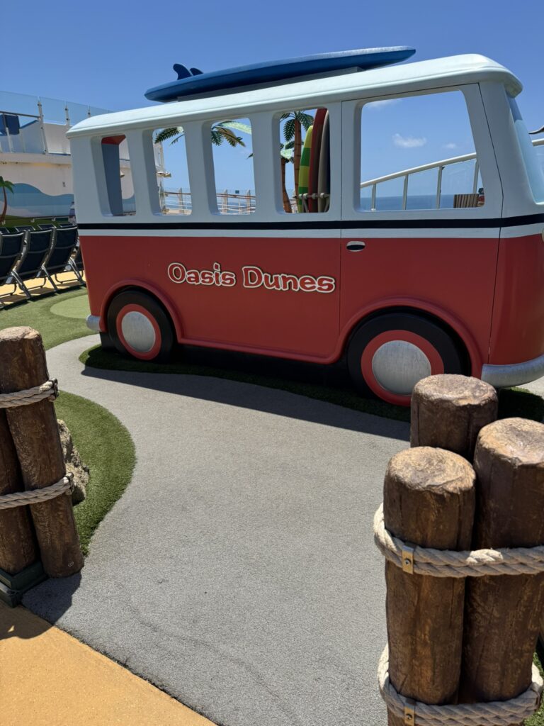 Retro‑style “Oasis Dunes” surf van display on a cruise ship deck, surrounded by beach‑themed décor and ocean views.