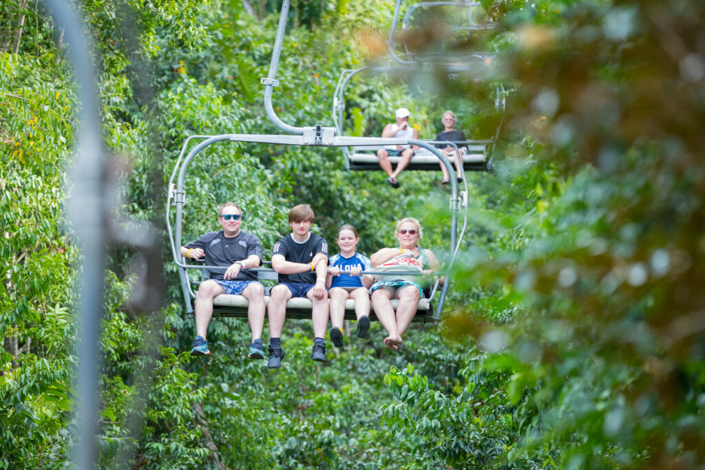 A group of tourists riding the SkyExplorer chairlift through the rainforest at Mystic Mountain in Ocho Rios, Jamaica, with views of the lush hills below.