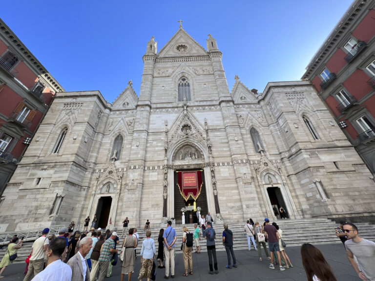 Exterior of Naples Cathedral with its Neo‑Gothic façade, pointed arches, and ornate stone detailing along Via Duomo