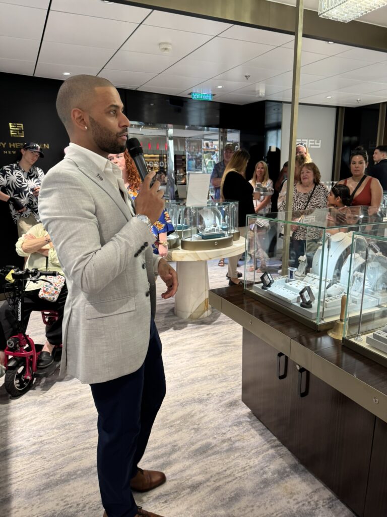Presenter speaking to guests during an onboard shopping event in the cruise ship’s jewelry store, with displays of rings and watches in the background.