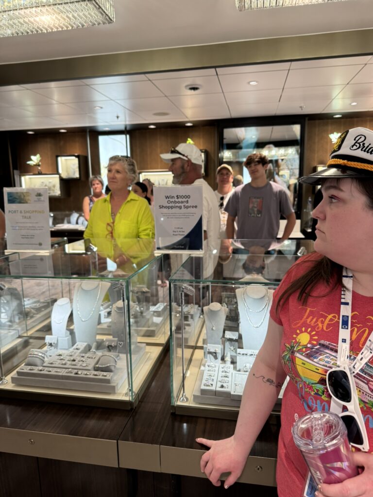 Guests gathered around jewelry displays during an onboard shopping event, with signs promoting port shopping talks and a chance to win an onboard shopping spree.