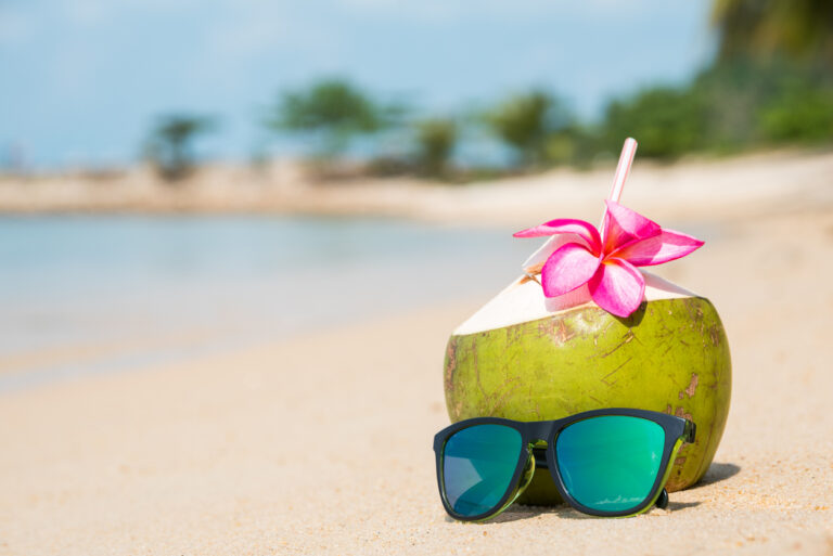 Coconut and sunglasses resting on a sandy tropical beach with bright turquoise water in the background, reflecting the relaxed, sunny atmosphere of a Caribbean shoreline.