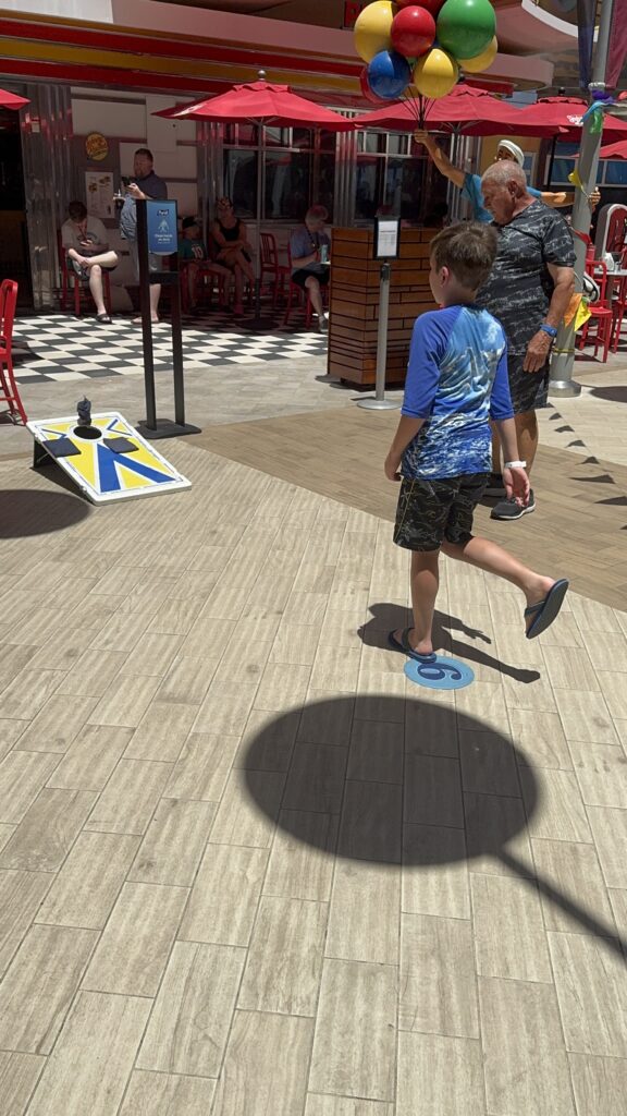 A child tosses a bean bag during a cornhole game in the Boardwalk area, highlighting one of the many free play spaces designed for little ones onboard.