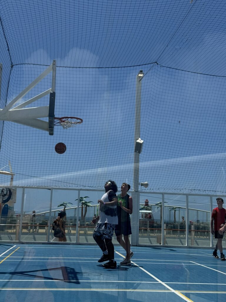 Guests playing basketball on the outdoor sports court of a Royal Caribbean ship with the ball mid‑air near the hoop