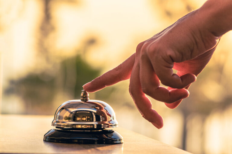 Hand ringing a polished service bell on a wooden counter, symbolizing concierge service and VIP attention Caption