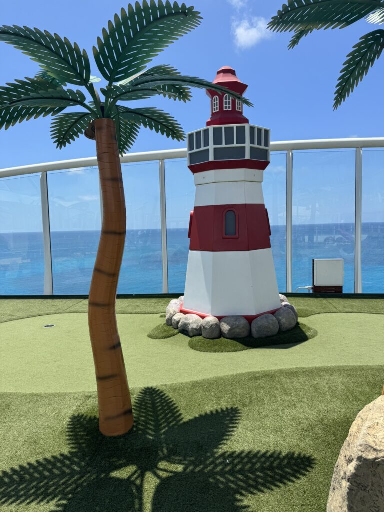 Mini golf course on a Royal Caribbean ship with a lighthouse feature, artificial palm trees, and ocean views through the deck’s glass panels