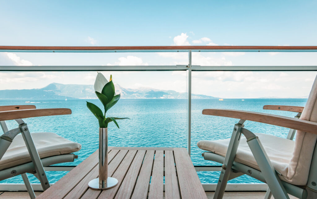 Elegant wooden table and chairs on a cruise ship balcony with a single flower and serene ocean and mountain views