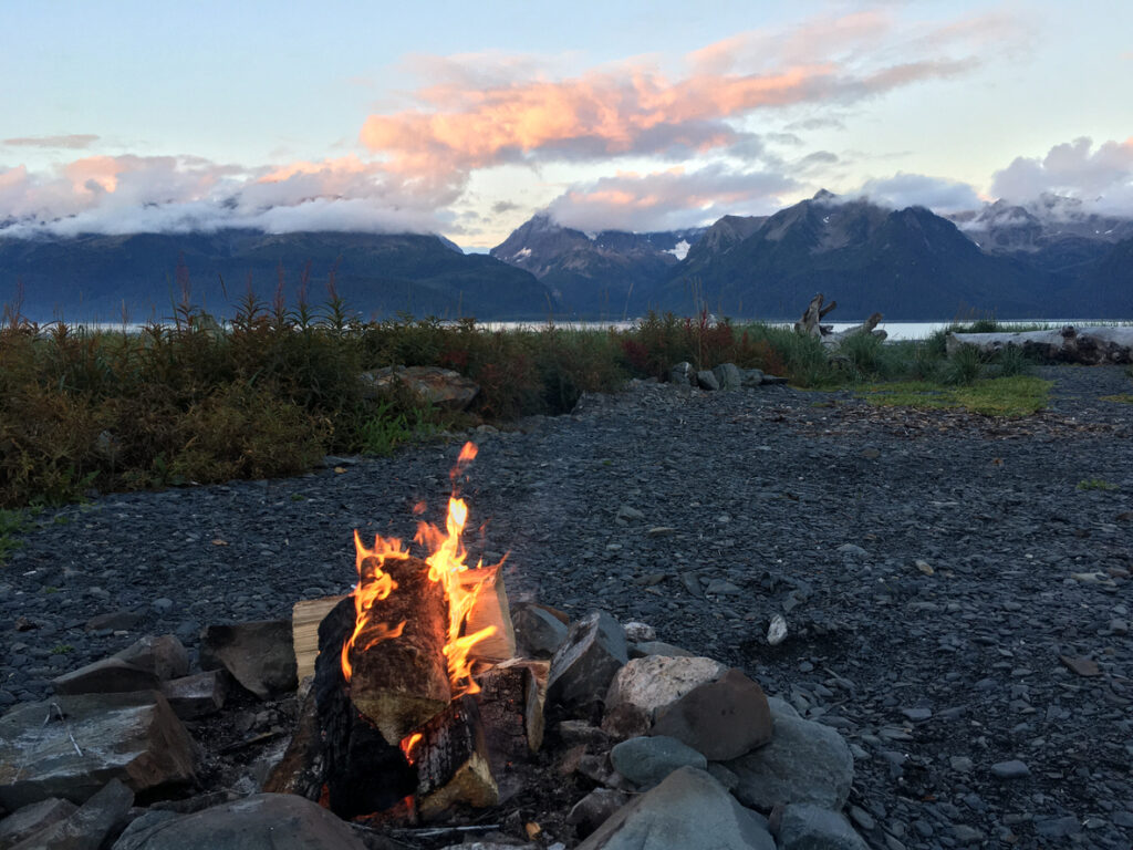 A glowing campfire on the rocky shoreline of Resurrection Bay in Seward, Alaska, with mountains rising across the water.