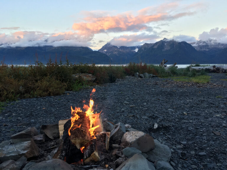 A glowing campfire on the rocky shoreline of Resurrection Bay in Seward, Alaska, with mountains rising across the water.
