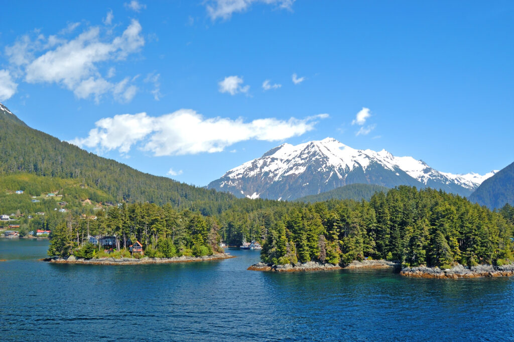 A forested Sitka coastline with houses along the shore, deep blue water, and snow‑capped mountains rising in the background under a clear sky.