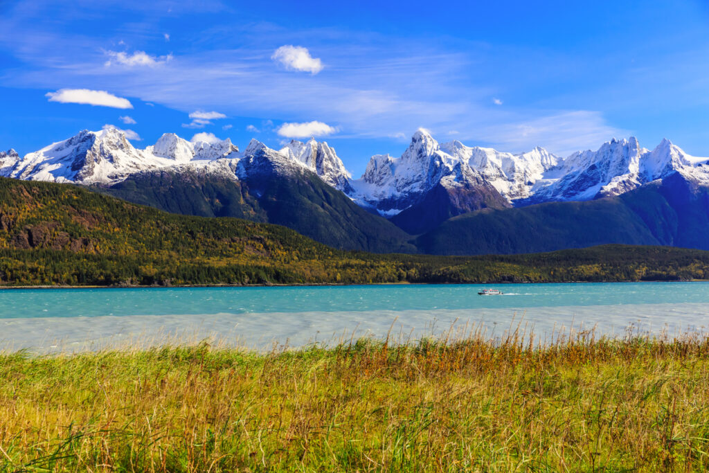 A turquoise lake with a small boat, surrounded by forested hills and towering snow‑capped mountains under a bright blue sky.
