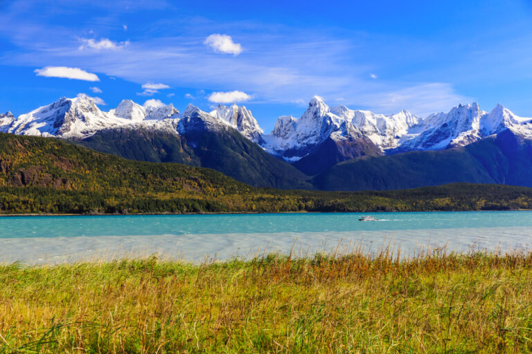 A turquoise lake with a small boat, surrounded by forested hills and towering snow‑capped mountains under a bright blue sky.
