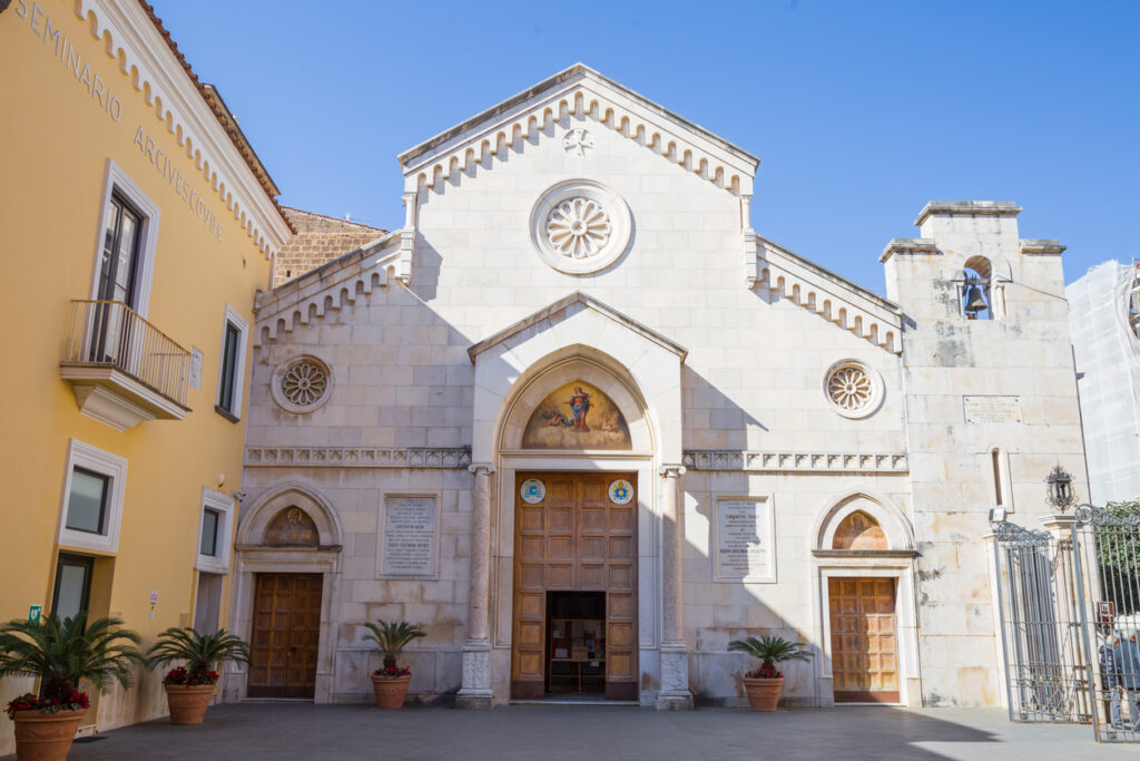 Exterior of Sorrento Cathedral with a Romanesque stone façade, arched doorway, rose windows, and a fresco above the entrance, with the bell tower and adjoining seminario building beside it