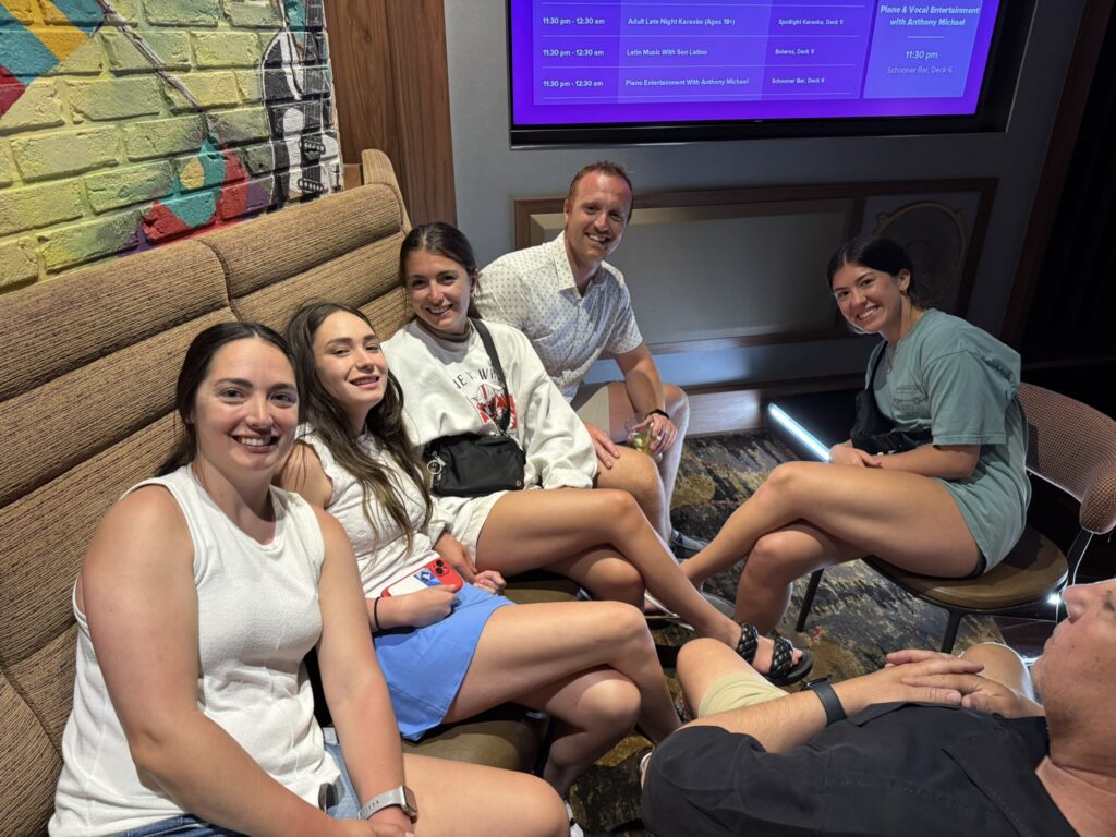 A group of people sit together in the Spotlight Karaoke lounge, playing a trivia game in a cozy seating area with the entertainment schedule glowing on the screen behind them.