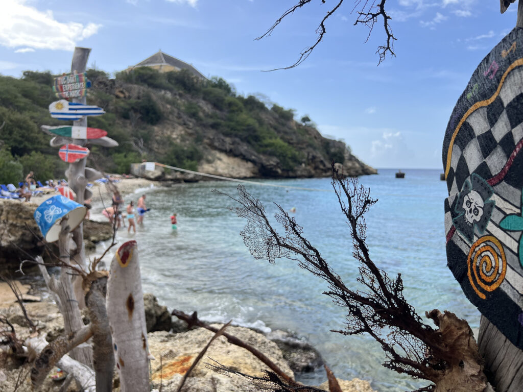 Close‑up of a branch and painted driftwood at Tugboat Beach in Curaçao with people relaxing and playing near the shoreline in the background