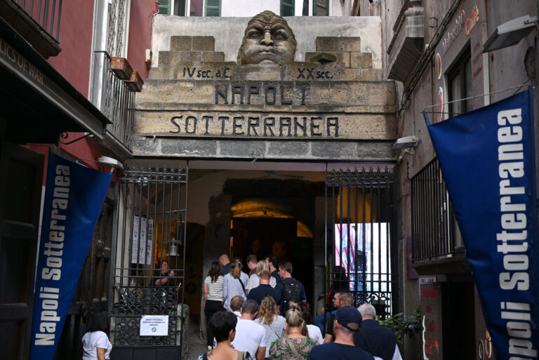 Entrance to Napoli Sotterranea with stone archway, carved face above the doorway, and visitors walking into the underground tour