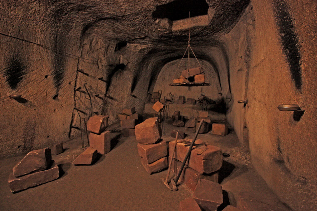 Underground stone chamber with large carved tuff blocks, metal tools, and a suspended platform used for historic stone‑cutting inside Napoli Sotterranea