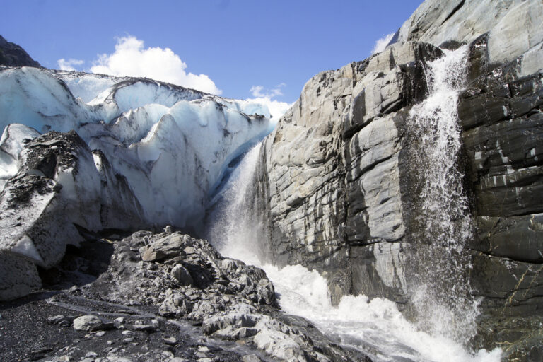 A blue‑tinted glacier beside a rocky cliff with a powerful waterfall cascading into white rapids under a bright sky.